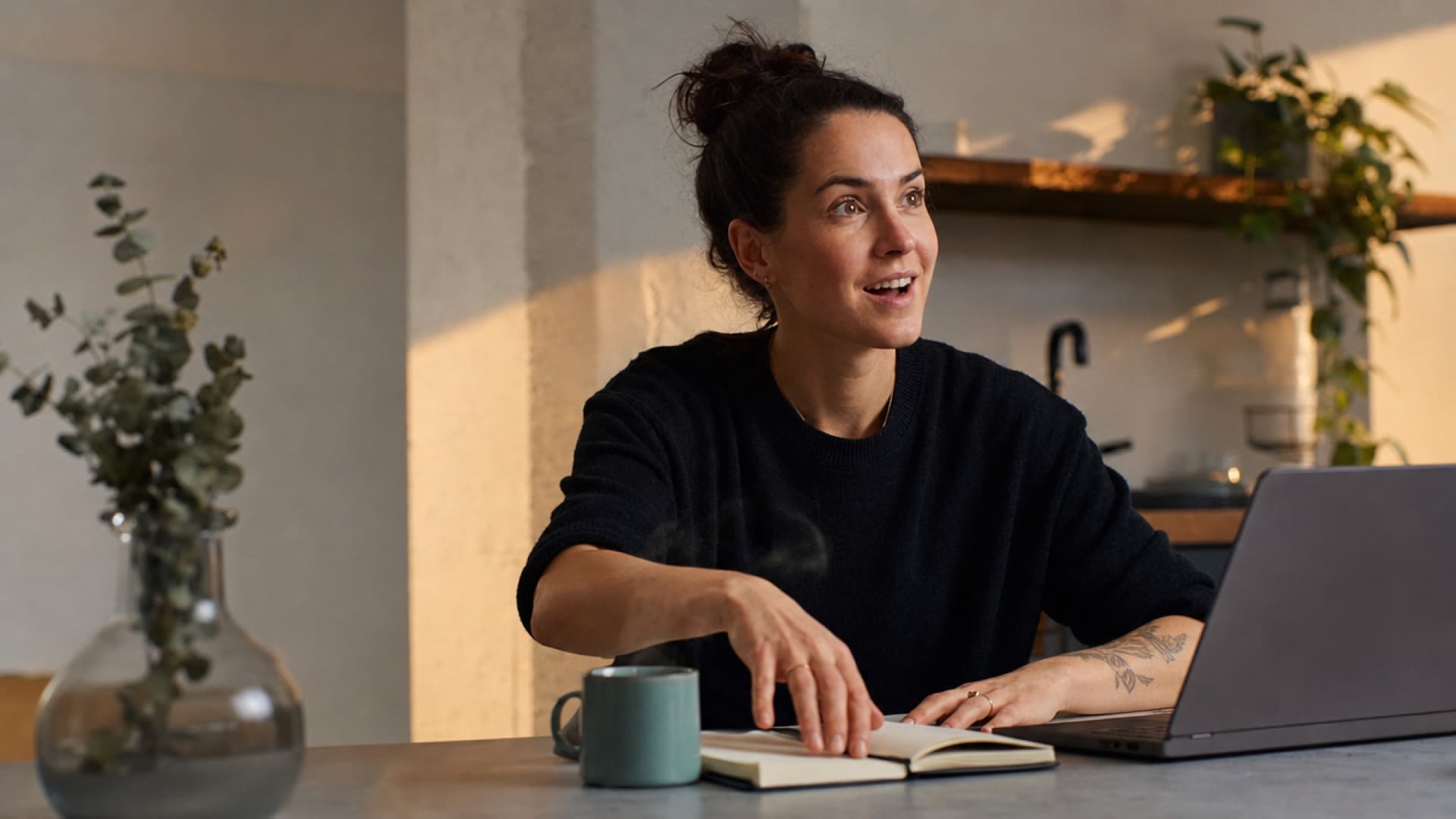 A woman at her kitchen table at dawn, smiling as she works on a laptop